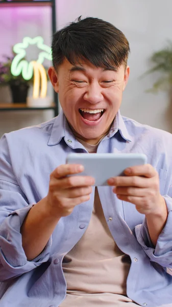 自宅でスマートフォンでレースやシューティングゲームゲームをプレイする心配アジアの勝者の男 リビングルームでソファに座ってドライブシミュレーターとスマートフォンのガジェットアプリを使用して中国の男 — ストック写真