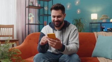 Kanepede oturan beyaz adam evde otururken cep telefonuyla gülümsüyor. Çevrimiçi akıllı telefon sosyal medya uygulamaları üzerinden mesajlaşan genç adam, rahatlama filmi izliyor