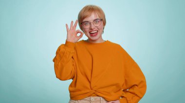 Kazaklı neşeli genç bir kadın kameraya onaylı geri bildirim jesti göstererek olumlu bir işaret veriyor. Mavi arka planda yalnız güzel bir kız. İnsanlar samimi duygular