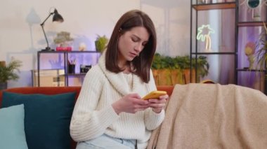 Hüzünlü kadın akıllı telefon daktilo kullanıyor, beklenmedik piyango sonuçlarını kaybediyor, kötü haber, servet kaybı, başarısızlık, teslim tarihi, virüs. Evdeki kanepede oturan genç kızın portresi.