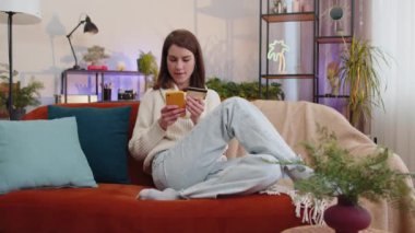 Para transfer ederken kredi kartı ve akıllı telefon kullanan genç bir kadın. İnternetten nakitsiz alışveriş yapıyor. Ev içinde yemek siparişi veriyor. Mutlu kız kanepede oturuyor