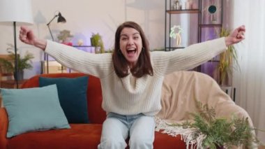 Happy young woman shouting, celebrating success, winning, goal achievement good news raising fists in gesture I did it, lottery luck at modern home apartment. Girl in living room sitting on couch