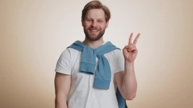 Genç beyaz adam zafer işareti gösteriyor, başarı ve kazanmayı umuyor, barış jesti yapıyor, iyimser bir ifadeyle gülümsüyor. Yakışıklı sakallı adam bej renkli stüdyoda tek başına izole edilmiş.