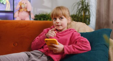 Young child kid girl texting messages on smartphone social media applications, watching relax movie, online shopping, play game. Female teen toddler uses mobile phone at home in room lying on sofa