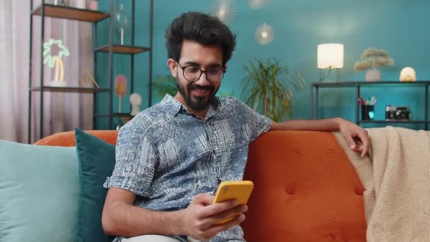 快乐的印地安人用手机打字说 发现大获全胜好消息庆祝彩票中奖做得奖手势 家里的印度教年轻人坐在沙发上 — 图库视频影像