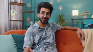 POV of indian man looking at camera, making video webcam conference call with friends or family, enjoying pleasant conversation at home room apartment. Hindu guy sits on sofa laughing, waving hello
