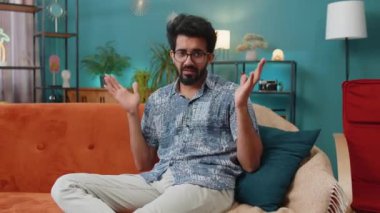 What. Why. Indian man raising hands in indignant expression asking reason of failure demonstrating disbelief irritation by troubles at modern home apartment. Hindu guy in living room sitting on sofa