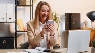 Happy amazed young businesswoman girl working on smartphone shocked by sudden victory game winning lottery jackpot achievemen good career news. Excited blonde woman freelancer sitting at home office