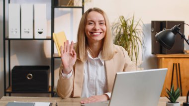 Young business woman working on laptop computer smiling friendly at camera and waving hands gesturing hello, hi, greeting or goodbye, welcoming with hospitable expression at home office workplace desk