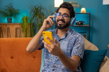 Happy overjoyed hindu indian man in wireless headphones relaxing sits on couch at home apartment choosing listening favorite energetic disco dancing music. People weekend daytime leisure activities