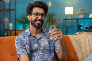 Susamış Hintli adam elinde bir bardak doğal su tutarak susuzluğu gidermek için bir yudum su içiriyor ve oturma odasında oturuyor. İyi yaşam alışkanlıkları olan Hindu adam, sağlıklı zayıflama, kilo verme konsepti
