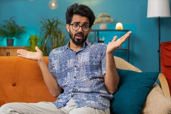 What. Why. Indian man raising hands in indignant expression asking reason of failure demonstrating disbelief irritation by troubles at modern home apartment. Hindu guy in living room sitting on sofa