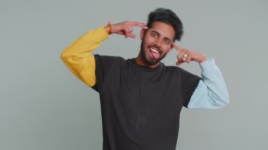 Komik, komik, genç Hintli bir adam şakacı, aptal yüz ifadeleri yapıyor, suratını buruşturuyor, aylak aylak dolanıyor, dil gösteriyor. Yakışıklı Hindu çılgın adam stüdyonun gri arka planında izole edilmiş.