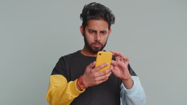 インド人の男性は 携帯電話の携帯電話の入力のブラウジングを使用すると 突然宝くじの結果 失敗に驚いて失う ハンサムなHindu男孤立した一人でグレーのスタジオの壁の背景に — ストック動画