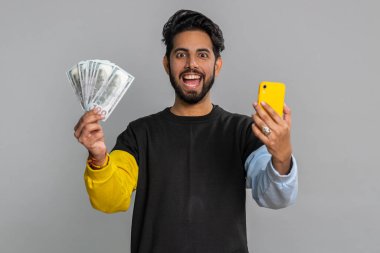 Akıllı telefona bakan Hintli bir adam içten bir sevinç gösterisi kazanır, para dolar banknotları, büyük ikramiye başarısı, gelir maaşı alır. Hintli adam gri arka planda izole edilmiş.
