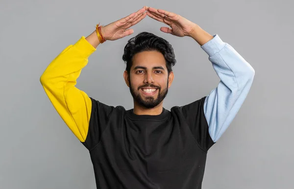 Young indian man playing childish catching up game, feeling in safe making roof above head with hands, insurance, security service. Bearded hindu guy isolated alone on gray studio background, indoors