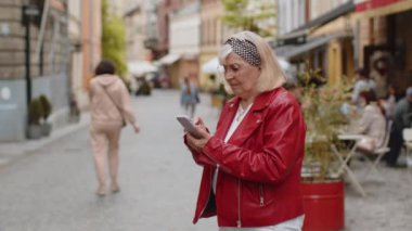 Gezgin son sınıf öğrencisi kadın akıllı telefon kullanıyor açık hava mobil navigatör uygulamasında haritada arama yapmak için kısa mesajlar yazıyor. Yaşlı bir büyükanne şehir sokağının önünden geçiyor.