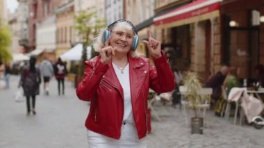 Kablosuz kulaklıklı mutlu son sınıf kadını favori enerjik müziği dinliyor, zaferi kutluyor. Yaşlı turist büyükanne şehir caddesinde yürüyor. Şehir yaşam tarzları açık havada
