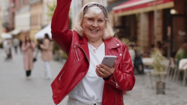 シニア女性は モバイルスマートフォンを使用しています受賞を祝う良いメッセージニュース 宝くじのジャックポットの勝利 プレゼントをオンライン 幸せな高齢者の祖母の屋外の都市の通りの背景に歩いてください ライフスタイル — ストック動画