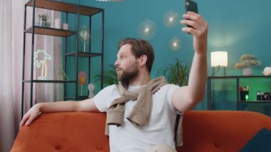Mutlu blogcu akıllı telefondan selfie çekiyor, abonelerle online video görüşmesi yapıyor, sosyal medya için hikayeler kaydediyor. Evdeki kanepede oturan beyaz adamın portresi.