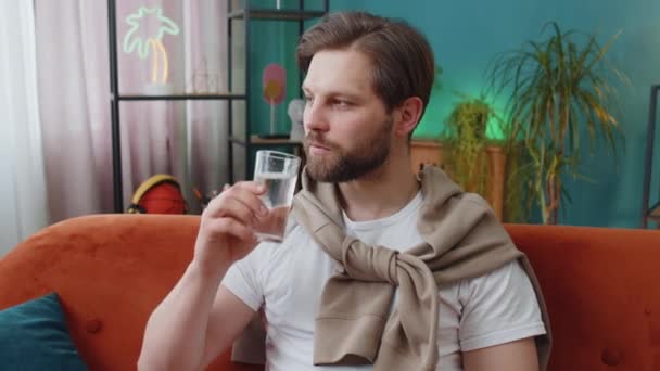 口渴的男人拿着一杯天然水 让小口小口喝水 防止脱水 坐在家里客厅里 白种人 生活习惯好 体形降 体重减轻观念 — 图库视频影像