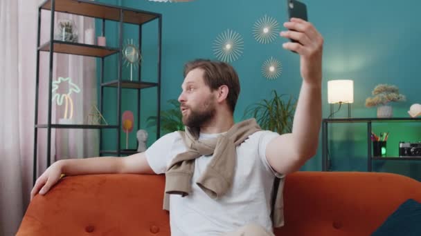 快乐的男人博主在智能手机上自拍 在网上与订户交流视频 为社交媒体记录故事 沙发上公寓客厅里白人男子的画像 — 图库视频影像