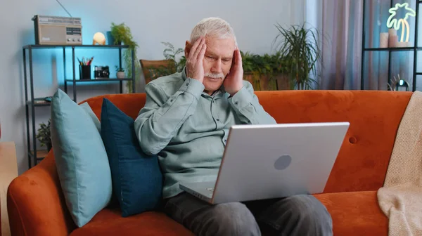 Yorgun büyükbaba dizüstü bilgisayar kullanıyor, baş ağrısı, tansiyon, migren, evde stres. Yaşlı adam defter üzerinde çalışıyor, mesajlar gönderiyor, online alışverişler yapıyor, film izliyor.