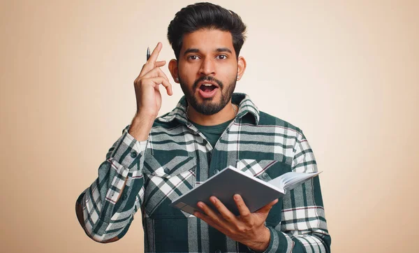 Thoughtful journalist indian man making notes, writing down thoughts with pen into notepad notebook diary, to do list, good idea. Handsome bearded hindu guy isolated alone on beige studio background