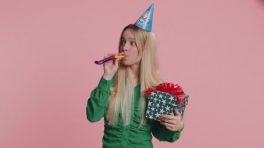 Doğum günü şapkası takan mutlu genç bir kadın zafer kutlaması için boru üflüyor. Yıldönümü partisinde heyecanlı şanslı kız pembe stüdyo arka planında kapalı kaldı. İnsan yaşam tarzı kavramı