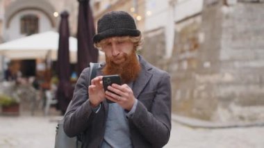 Açık hava mobil navigatör uygulamasında haritada bir yol bulmak için akıllı telefon kullanarak mesajlar yazan sakallı bir gencin portresi. Şehir sokaklarında yürüyen kızıl saçlı adam. Kasaba yaşam tarzları