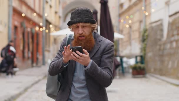 年轻的大胡子男人用智能手机打字浏览 输了意外的彩票结果 坏消息 红头发的家伙走在城市街道的室外 城镇生活方式 — 图库视频影像
