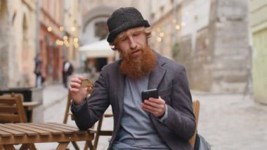 Genç adam para transfer ederken kredi kartı akıllı telefonu kullanıyor, online alışveriş yapıyor, yemek siparişi veriyor, otel odası ayırtıyor. Şehir caddesindeki kafede oturan kızıl saçlı adam.