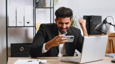 Indian businessman playing on smartphone exciting mobile application celebrate winning racing or shooter game at office desk workplace. Professional manager freelancer man enjoy good work of new app