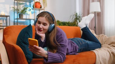 Portrait of happy overjoyed woman in wireless headphones relaxing sits on couch at home apartment choosing listening favorite energetic disco dancing music. People weekend daytime leisure activities