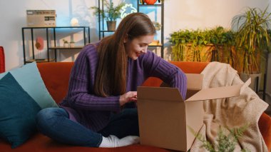 Evdeki kanepede oturan mutlu, genç bir kadın teslimat paketini açıyor. Gülümseyen tatmin olmuş kız alışverişci internet müşterisi karton kutu açıyor hızlı posta yoluyla satın alma hediyesi alıyor