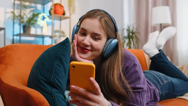 Portrait of happy overjoyed woman in wireless headphones relaxing sits on couch at home apartment choosing listening favorite energetic disco dancing music. People weekend daytime leisure activities