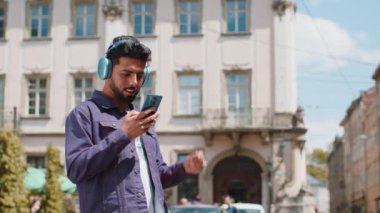 Kablosuz kulaklıklı, neşeli, neşeli genç Hintli adam dışarıda akıllı telefon dansında en sevilen disko rock 'n roll müziğini dinliyor. Şehir caddesinde yürüyen sakallı adam.