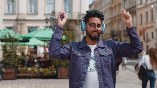 お気に入りの精力的な音楽を聞いて ワイヤレスヘッドフォンで幸せな喜びの若いインド人男性は 勝利を祝います ひげを生やした人観光客の旅行者の都市部の街を歩く 町のライフスタイル屋外 — ストック動画