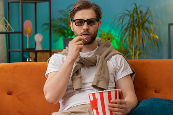 Excited young man in 3D glasses sits on sofa eating popcorn snacks and watching interesting TV serial sport game, film, online social media movie content at home apartment. Guy enjoying entertainment