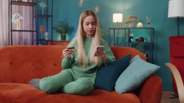 Beyaz bir kadın kredi kartı ve akıllı telefon kullanarak para transfer ediyor, kapalı bir evde alışveriş nakitsiz yemek siparişi veriyor. Mutlu kız kanepede oturuyor