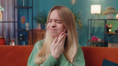 Diş sorunları. Yanağa dokunan genç bir kadın, korkunç bir ifadeyle gözlerini kapatan diş ağrısı, hassas dişler, çürükler. Evdeki genç kız turuncu koltukta oturuyor.