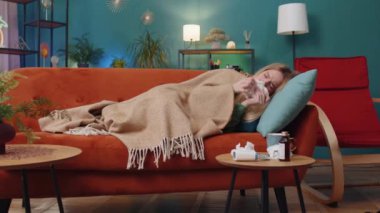 Evdeki oturma odasındaki kanepede yatan soğuk algınlığı ya da alerjiden muzdarip bir kadın. Kafasında havlu olan hasta çocuk hapşırıyor, peçeteye sümük döküyor. Coronavirus karantina salgını