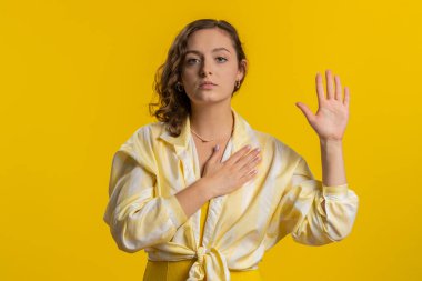 Dürüst olacağıma yemin ederim. İçten, sorumluluk sahibi genç bir kadın yemin etmek için elini kaldırıyor, dürüst olacağına ve doğruyu söyleyeceğine söz veriyor, elini göğsüne koyuyor. Sarı stüdyo arka planında izole edilmiş bir kız. Yaşam Tarzları