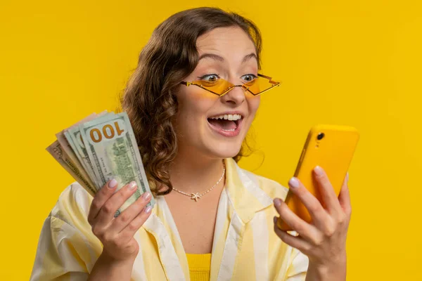 スマートフォンを探している若い女性は 成功宝くじ運を祝う お金ドルの現金紙幣を受け取り 誠実に勝利を喜んで表示されます 黄色のスタジオの背景に孤立した女の子 ライフスタイル — ストック写真