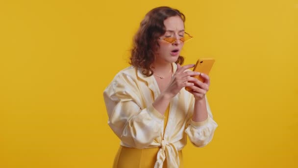 女性は 携帯電話のスマートフォンのタイプブラウジングを使用し 突然の宝くじの結果 失敗に驚いて失う かなりブルネットの女の子一人で黄色のスタジオの背景に — ストック動画