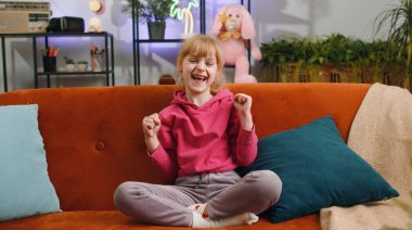 Çocuk kız bağırıyor, başarıyı kutluyor, kazanıyor, gol başarısı, iyi haber, el hareketleriyle yumruk kaldırıyor. Başardım, piyango şansı. Genç bir kız evde oyun oynuyor oturma odasında kanepede oturuyor.