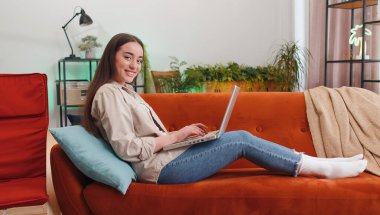 Woman freelancer opens laptop, starts working education on notebook, sends messages, makes online purchases, watching movies at home living room apartment. Portrait of girl sitting on couch. Lifestyle