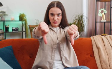 Dislike. Portrait of upset woman showing thumbs down sign gesture, expressing discontent, disapproval dissatisfied bad work at modern home apartment indoors. Displeased girl in living room on sofa