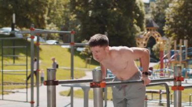 Atletik beyaz, üstsüz, kaslı bir adam paralel barlarda dalış yapıyor, trisepsleri eğitiyor. Çocuk parkındaki genç üstsüz adam. Spor sağlık programı, antrenman. Güç motivasyonu. Açık spor salonu.