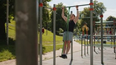 Spor giyimli atletik bir adam elastik genişleticinin yardımıyla barfiks egzersizi yapıyor. Çocuk parkında sırt kaslarını şişiren genç adam. Spor sağlığı rutin egzersizi. Vücut geliştirme
